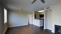 This single-floor residence features an open-concept layout that seamlessly connects the living area to the kitchen. The living space is finished with rich, dark hardwood flooring that extends throughout, creating a warm and cohesive aesthetic. Neutral gray walls provide a clean, modern backdrop, while white baseboards offer a crisp contrast.

The living area is equipped with a ceiling fan for year-round comfort and a track lighting fixture mounted on the wall, allowing for flexible, directional illumination. Multiple electrical outlets are conveniently positioned along the walls.

The kitchen is partially separated by a raised breakfast bar with a dark countertop, providing both a casual dining option and a natural division between spaces. Dark espresso-toned cabinetry lines the kitchen walls, complemented by black appliances including a microwave. Overhead fluorescent lighting ensures the kitchen is well-lit for meal preparation.

A doorway visible from the main living area leads to additional rooms beyond, suggesting a functional floor plan with private spaces accessible from the central living zone. The overall ambiance is modern and understated, with a neutral palette suited to a variety of interior styles.