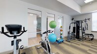 This fitness center is designed to support a well-rounded exercise routine in a clean, modern environment. The space features light wood-style flooring and crisp white walls that contribute to a bright, open atmosphere. Natural and artificial lighting is distributed throughout, keeping the area feeling fresh and energizing.

The main workout area is equipped with a variety of fitness tools, including a stationary spin bike, a multi-station weight machine, and a medicine ball rack stocked with an assortment of weighted and stability balls. Exercise mats are also available, making the space suitable for flexibility and floor-based training as well.

Large glass-paneled doors and interior windows connect the main gym floor to an adjacent enclosed room, which appears to be a dedicated space for stretching, yoga, or small-group exercise. This separation allows for versatile use of the facility while maintaining a sense of openness throughout.

The overall layout is thoughtfully organized, with equipment arranged to allow comfortable movement between stations. The single-floor layout ensures easy accessibility, and the contemporary design aesthetic creates a motivating environment for users of all fitness levels.