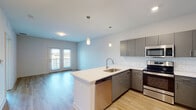This thoughtfully designed single-floor residence showcases a contemporary open-concept layout that seamlessly connects the kitchen and living areas. The kitchen is a standout feature, equipped with sleek gray flat-front cabinetry, a classic white subway tile backsplash, and a full suite of stainless steel appliances including a refrigerator, range, microwave, and dishwasher. A peninsula with a white quartz countertop and an undermount sink anchors the space, illuminated by stylish pendant lighting overhead. Light wood-look flooring flows throughout the main living area, enhancing the bright, airy ambiance created by the neutral white walls. The living area leads to a set of French doors that open onto a balcony, allowing natural light to pour in and offering outdoor views. The bedroom is finished with plush carpeting, a ceiling fan, and a window framing exterior greenery. It connects to a generously sized walk-in closet with wire shelving for ample storage. The bathroom features a wide white quartz vanity, a large mirror, and a subway-tiled combination tub and shower. Throughout the space, clean lines, modern finishes, and a cohesive neutral palette contribute to a polished, move-in-ready aesthetic.