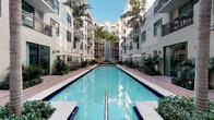 This residential community showcases a striking contemporary design built around a central outdoor courtyard that serves as its defining feature. At the heart of the property lies a generously sized lap pool with crystal-clear blue water, framed symmetrically by the surrounding multi-story buildings. The pool area is thoughtfully appointed with lounge chairs, shading umbrellas, and tropical landscaping, including tall swaying palm trees that lend the space a distinctly resort-like atmosphere. The architecture features a clean, modern aesthetic with a bright white exterior, large windows, and private balconies that overlook the courtyard below. The open-sky design of the courtyard allows abundant natural light to flood the space throughout the day, creating a warm and inviting ambiance. Brick-paved walkways border the pool on both sides, providing well-defined pathways and gathering areas for residents. The lush greenery and carefully arranged plantings throughout the courtyard add a natural, vibrant contrast to the building's crisp exterior. Overall, the community blends upscale architectural design with resort-inspired outdoor living, offering a refined and comfortable environment centered around relaxation and community enjoyment.
