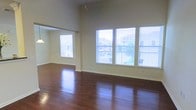 This two-floor space showcases an impressive open layout defined by soaring ceilings that lend a dramatic sense of volume and openness. The main living area is anchored by rich, dark hardwood flooring that runs throughout, reflecting light and adding warmth to the overall aesthetic. Large windows dressed with horizontal blinds line the walls, inviting generous amounts of natural light and offering views of the surrounding neighborhood and greenery outside. The high ceiling height creates an airy, expansive atmosphere that makes the main living space feel particularly grand. A pendant light fixture hangs from above, adding a subtle design accent that complements the clean, neutral wall tones. The open-plan layout allows for flexible furniture arrangements and seamless flow between areas. An adjacent room or alcove is visible beyond a wide architectural opening, further enhancing the sense of connected, spacious living. The overall ambiance is bright, airy, and welcoming, with a classic yet timeless interior character well-suited for a variety of living arrangements. The combination of elevated ceilings, polished hardwood floors, and plentiful windows makes this a standout space full of natural charm and functional versatility across both its floors.
