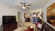 This single-story residence at 1800 Links Blvd in Tuscaloosa, Alabama, offers an open-concept floor plan that seamlessly connects the main living areas. The living room serves as the central gathering space, featuring light-toned carpet flooring and a ceiling fan with integrated lighting that provides both comfort and illumination. A large flat-screen television is positioned on a rich, dark wood media console, complemented by comfortable seating including an upholstered armchair and sofa accented with decorative throw pillows in warm reds and neutrals.

The open layout flows directly into a dedicated dining area, which accommodates a full dining table and chairs, making it well-suited for everyday meals or entertaining. Adjacent to the dining space, the kitchen is equipped with white cabinetry, standard appliances including a refrigerator, and a countertop bar with bar stool seating — adding both functionality and a casual dining option. The kitchen benefits from its own ceiling fan and overhead lighting. Throughout the space, the warm color palette, soft flooring, and connected layout create a cohesive and welcoming atmosphere suited for comfortable daily living.