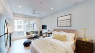 This thoughtfully designed single-floor studio offers a seamless open-concept layout that combines sleeping, living, and cooking areas into one cohesive space. Light hardwood flooring runs throughout, complementing the soft, neutral color palette of pale blues, whites, and warm beiges that give the interior a calm and airy feel.

The main living and sleeping area is generously proportioned, featuring recessed lighting, a ceiling fan, and large windows that invite abundant natural light and views of the surrounding urban streetscape. A dedicated lounge nook with a sofa, glass coffee table, wall-mounted TV, and a compact desk provides distinct functional zones within the open floor plan.

The kitchen is fully equipped with stainless steel appliances — including a refrigerator, dishwasher, oven, and microwave — set against crisp white cabinetry with gold-toned hardware and a striking marble-style backsplash. Under-cabinet lighting and white quartz countertops add a polished, modern touch.

The bathroom is finished with warm travertine-style wall and floor tiles, a floating white vanity with gold hardware, and a glass-enclosed walk-in shower. Framed artwork adds a refined accent to the space.

The entry hallway features light walls, natural wood flooring, and tasteful decorative accents, creating a welcoming transition into the main living space.