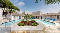 This outdoor amenity space is designed around a generously sized, multi-section swimming pool that serves as the central focal point of the property. The pool is divided into distinct areas, including a main swimming section with clear, blue-tinted water and a separate shallow wading zone lined with cobalt blue tile accents. A stone-faced waterfall feature connects the two pool sections, adding both visual appeal and ambient sound to the environment.

A standout architectural element is the raised central planter island situated within the pool area, landscaped with a palm tree and vibrant seasonal flowers, creating a tropical aesthetic. The pool surround is finished with natural stone coping and a combination of concrete and paved decking, providing ample space for lounge chairs arranged throughout.

The buildings flanking the pool area feature a neutral gray lap siding exterior with pitched rooflines and accented blue entry doors. A covered outdoor patio area is attached to one of the buildings, offering shade and shelter. It is furnished with blue upholstered seating, a metal coffee table, and a ceiling fan overhead, creating a comfortable space for lounging. Mature trees provide additional shade around the perimeter, and a fenced boundary ensures privacy throughout the outdoor space.