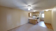 This single-floor space offers a straightforward, functional layout centered around an open-concept living and kitchen area. The living space features neutral carpeting and a ceiling fan with a light fixture, contributing to a comfortable and well-lit atmosphere. A textured ceiling adds subtle character throughout the main area.

The kitchen is positioned at the heart of the space, defined by a peninsula with a built-in sink that serves as a natural dividing point between the cooking and living areas. Warm wood-tone cabinetry lines the upper walls of the kitchen, providing ample storage. White appliances, including a refrigerator and a gas range with overhead ventilation, are neatly arranged along the kitchen wall. A window near the kitchen allows natural light to filter in, brightening the space.

The flooring transitions from carpet in the living area to a hard surface in the kitchen and adjacent zones, offering both comfort and practicality depending on the function of each area. The overall color palette is neutral and understated, with beige and warm tan tones dominating the walls and surfaces.

The layout is efficient and easy to navigate, making it well-suited for everyday living. The open sightlines between the kitchen and living area create a sense of connectivity and spaciousness throughout this compact, single-level space.