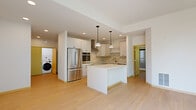 This contemporary single-floor residence showcases a clean, minimalist aesthetic throughout, with light hardwood flooring and crisp white walls creating a cohesive and airy atmosphere. The kitchen serves as a central highlight, featuring white shaker cabinetry with matte black hardware, a striking herringbone marble-look backsplash, stainless steel appliances including a French door refrigerator, double wall ovens, a cooktop with a stainless range hood, and a built-in microwave. A central island with a white quartz countertop offers additional prep space, while pendant lighting adds a refined touch overhead. An in-unit washer and dryer are conveniently accessible from the kitchen area. The bedrooms are generously proportioned, each filled with abundant natural light through large windows that frame lush tree canopy views. Ceiling fans are present in multiple rooms for added comfort. One bedroom connects directly to a well-lit bathroom with a wide vanity. A standout feature is the spacious private terrace accessible via full-height sliding glass doors, offering an open-air retreat complete with a gas grill. The overall design balances modern functionality with light, nature-connected interiors throughout.