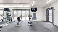 Located at 304 Madison Ave in Boone, NC, this fitness center offers a spacious, single-floor workout facility designed to accommodate a wide range of exercise needs. The open floor plan provides ample room for movement and equipment use, all laid out across durable, dark rubber-tile flooring ideal for high-traffic athletic use.

The space features a coffered white ceiling with bright fluorescent lighting, ensuring strong, even illumination throughout. One wall is finished in a bold blue-painted concrete block, adorned with large wall-mounted mirrors that expand the sense of space and allow users to monitor their form during workouts. A wall-mounted flat-screen television is positioned for visibility across the floor.

Large sliding glass doors and framed windows along one side of the facility flood the interior with natural light and provide views of the surrounding outdoor environment. A mini-split HVAC unit ensures climate control year-round.

Equipment includes multiple treadmills, stationary and recumbent bikes, a multi-function cable resistance machine, a Jones machine, free weight racks with dumbbells and barbells, a weight bench, and additional functional training accessories such as medicine balls. The facility is well-organized, allowing users to transition easily between cardio, strength, and functional training zones within a single, cohesive space.