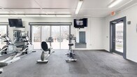Located at 304 Madison Ave in Boone, NC, this fitness center offers a spacious, single-floor workout facility designed to accommodate a wide range of exercise needs. The open floor plan provides ample room for movement and equipment use, all laid out across durable, dark rubber-tile flooring ideal for high-traffic athletic use.

The space features a coffered white ceiling with bright fluorescent lighting, ensuring strong, even illumination throughout. One wall is finished in a bold blue-painted concrete block, adorned with large wall-mounted mirrors that expand the sense of space and allow users to monitor their form during workouts. A wall-mounted flat-screen television is positioned for visibility across the floor.

Large sliding glass doors and framed windows along one side of the facility flood the interior with natural light and provide views of the surrounding outdoor environment. A mini-split HVAC unit ensures climate control year-round.

Equipment includes multiple treadmills, stationary and recumbent bikes, a multi-function cable resistance machine, a Jones machine, free weight racks with dumbbells and barbells, a weight bench, and additional functional training accessories such as medicine balls. The facility is well-organized, allowing users to transition easily between cardio, strength, and functional training zones within a single, cohesive space.