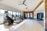 Located at 4418 Oat Grass Circle in Charlotte, NC, this well-appointed fitness center offers a thoughtfully designed single-floor layout that balances functionality with contemporary style. The space is defined by an open floor plan with light hardwood-style flooring throughout the cardio zone, transitioning to rubberized flooring in the strength and free-weight areas. Large floor-to-ceiling windows line one wall, flooding the interior with abundant natural light while offering outdoor views of the surrounding landscape. Exposed wooden beams and a striking exposed brick accent wall add warmth and industrial character to the otherwise clean, modern aesthetic. Ceiling fans are positioned throughout to maintain comfortable airflow. The cardio section is equipped with a lineup of treadmills, a stair climber, and a stationary bike, while the strength zone features a multi-use cable and rack system, a free-weight bench with barbell, and a well-stocked dumbbell rack. Medicine balls, resistance tools, and a full-length mirror wall complete the strength area. A water fountain station is conveniently integrated into the space. The combination of modern equipment, architectural detail, and generous natural light makes this fitness center an inviting and fully functional amenity.