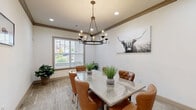 Located at 1110 Ballpark Ln in Lawrenceville, GA, this refined dining room offers a cohesive and inviting atmosphere with thoughtful design details throughout. The space is anchored by a rectangular glass-top dining table surrounded by tufted cognac leather chairs with nailhead trim, providing both elegance and comfort. Overhead, a wrought-iron candle-style chandelier serves as a striking focal point, complementing the warm wood-tone crown molding that frames the tray ceiling. Recessed lighting adds a soft, ambient glow throughout.

The flooring features wood-look tile in neutral grey and tan tones, contributing to the room's relaxed yet refined aesthetic. A large window fitted with horizontal blinds allows natural light to filter in, offering a view of the surrounding greenery. Potted plants placed throughout the room add a fresh, natural touch.

The walls are adorned with Western-inspired artwork, including a bold highland cow canvas print and a framed bison landscape, reinforcing a distinct rustic-modern character. A multi-pane French door with a dark frame connects the dining room to an adjacent space, adding architectural interest. A wall-mounted TV and a wooden credenza complete the room, making it as functional as it is visually appealing.