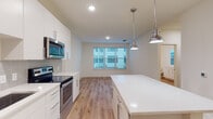 This contemporary single-floor residence showcases a clean, modern aesthetic with an open-concept layout that connects the kitchen and living areas seamlessly. The kitchen is a standout feature, equipped with a full suite of stainless steel appliances — including a French door refrigerator, electric range, built-in microwave, and dishwasher. Crisp white shaker-style cabinetry with sleek chrome hardware lines the walls, complemented by a subtle gray tile backsplash and bright white quartz countertops. A generously sized central island provides additional workspace and storage, while polished chrome pendant lights add a stylish finishing touch overhead. Recessed lighting illuminates the space throughout. Light-toned hardwood flooring flows consistently across all rooms, enhancing the bright and airy feel of the interior. The bedroom offers a comfortable layout with a ceiling fan, natural light from a large window with exterior views, and ample wall space. The bathroom features classic white subway tile surrounding a soaking tub and shower combination, a window for natural light, and continued hardwood flooring. Neutral wall tones and clean lines throughout the space contribute to a fresh, move-in-ready atmosphere.