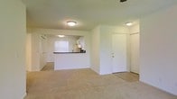 This single-floor residence offers a straightforward and functional layout designed for comfortable everyday living. The main living area is open and spacious, featuring neutral-toned carpet flooring and clean white walls that create a bright, airy atmosphere. Flush-mount ceiling light fixtures illuminate the space evenly throughout.

A defining architectural feature is the half-wall pass-through that connects the living area to the kitchen, promoting an open-concept feel while maintaining a subtle visual separation between spaces. The kitchen is positioned toward the rear and includes white cabinetry, dark countertops, and a vertical blind-covered window that allows natural light to filter in. Appliances are integrated into the kitchen layout, providing practical functionality.

The entry area features a white paneled door with a closet nearby, offering convenient storage upon arrival. Additional doors visible from the main living space suggest access to further rooms or storage areas, contributing to the home's overall utility.

The consistent use of white throughout the walls, trim, and doors, paired with the light carpeting, gives the interior a clean and neutral aesthetic that serves as a versatile backdrop for a variety of furnishing styles. The single-floor design ensures easy accessibility and a cohesive flow throughout the entire living space.