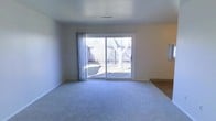 This single-floor space features a generously sized main living area finished with plush wall-to-wall carpeting in a neutral grey tone, contributing to a clean and understated aesthetic. The room is brightened by a large sliding glass door dressed with sheer curtains, which floods the interior with natural light and provides a direct view of the fenced backyard. Outside, mature deciduous trees and wooden privacy fencing frame the outdoor area, offering a sense of seclusion. The smooth white walls and white ceiling create a bright, open atmosphere, while recessed HVAC venting is visible overhead, indicating a central climate control system. A transition to what appears to be hard surface flooring is visible at the edge of the room, suggesting adjacent spaces with different finishes. The overall ambiance is calm and neutral, providing a versatile canvas well-suited to a variety of interior arrangements and décor styles.