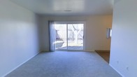 This single-floor space features a generously sized main living area finished with plush wall-to-wall carpeting in a neutral grey tone, contributing to a clean and understated aesthetic. The room is brightened by a large sliding glass door dressed with sheer curtains, which floods the interior with natural light and provides a direct view of the fenced backyard. Outside, mature deciduous trees and wooden privacy fencing frame the outdoor area, offering a sense of seclusion. The smooth white walls and white ceiling create a bright, open atmosphere, while recessed HVAC venting is visible overhead, indicating a central climate control system. A transition to what appears to be hard surface flooring is visible at the edge of the room, suggesting adjacent spaces with different finishes. The overall ambiance is calm and neutral, providing a versatile canvas well-suited to a variety of interior arrangements and décor styles.