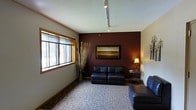 This single-floor space features a thoughtfully designed interior that balances comfort and functionality. The main seating area draws attention with a rich, deep burgundy accent wall that serves as a striking focal point, complemented by neutral-toned surrounding walls. Track lighting runs along the ceiling, providing adjustable, even illumination throughout the room, while a textured ceiling adds subtle architectural interest.

The space is furnished with dark navy leather seating, including a two-cushion sofa and an additional chair, arranged to encourage a relaxed, welcoming environment. A framed landscape artwork hangs on the accent wall, enhancing the room's visual appeal. A small side table with a warm table lamp adds a soft, ambient glow that balances the overhead lighting.

Natural light enters through a wide, wood-trimmed window fitted with blinds, offering both brightness and privacy control. The floor is covered in a patterned area rug over carpeting, adding warmth and texture underfoot. Decorative dried branches in a tall vase contribute an organic, natural element to the overall aesthetic. The space presents a polished yet comfortable ambiance well-suited for receiving visitors or quiet relaxation.