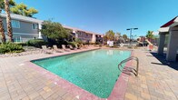 This inviting outdoor amenity space is thoughtfully designed for both relaxation and social gatherings in a classic desert setting. At the heart of the area lies a generously sized rectangular swimming pool with clear turquoise water, surrounded by a paved brick deck that provides ample room for lounging. A row of sling-style chaise lounges lines the poolside, offering residents comfortable spots to soak up the abundant sunshine under open blue skies.

A covered pavilion with a distinctive red metal roof and sturdy brick columns provides a shaded retreat, furnished with lounge chairs for those seeking a break from the sun. A separate covered picnic area features a red-framed corrugated metal roof and classic wooden picnic tables, creating an ideal setting for casual outdoor dining and gatherings.

The space is equipped with multiple built-in BBQ grills — including a substantial stucco-and-tile outdoor grill station — making it well-suited for cookouts and community events. Desert-appropriate landscaping, including palm trees, ornamental shrubs, and decorative rock ground cover, frames the area and enhances its Southwest character. A wrought-iron perimeter fence encloses the space, providing both security and open views of the surrounding greenery and neighborhood beyond.