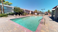 This inviting outdoor amenity space is thoughtfully designed for both relaxation and social gatherings in a classic desert setting. At the heart of the area lies a generously sized rectangular swimming pool with clear turquoise water, surrounded by a paved brick deck that provides ample room for lounging. A row of sling-style chaise lounges lines the poolside, offering residents comfortable spots to soak up the abundant sunshine under open blue skies.

A covered pavilion with a distinctive red metal roof and sturdy brick columns provides a shaded retreat, furnished with lounge chairs for those seeking a break from the sun. A separate covered picnic area features a red-framed corrugated metal roof and classic wooden picnic tables, creating an ideal setting for casual outdoor dining and gatherings.

The space is equipped with multiple built-in BBQ grills — including a substantial stucco-and-tile outdoor grill station — making it well-suited for cookouts and community events. Desert-appropriate landscaping, including palm trees, ornamental shrubs, and decorative rock ground cover, frames the area and enhances its Southwest character. A wrought-iron perimeter fence encloses the space, providing both security and open views of the surrounding greenery and neighborhood beyond.
