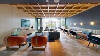 The Alta Botbero amenity lounge is a thoughtfully designed, single-floor communal space that seamlessly blends refined aesthetics with everyday functionality. A striking coffered wood ceiling serves as the architectural centerpiece, adding warmth and texture throughout the open-plan interior, while herringbone-patterned hardwood flooring grounds the space with understated elegance.

The main lounge area is generously appointed with a curated mix of seating — including plush gray sofas, distinctive rust-colored velvet armchairs, and sculptural wicker accent chairs — all arranged around a central coffee table atop a deep teal area rug. A wall-mounted flat-screen television is integrated into a sleek, full-height built-in cabinetry unit, creating a media focal point ideal for both relaxed lounging and social gatherings.

A glass-partitioned kitchenette runs along one side of the space, outfitted with light cabinetry, countertops, and beverage stations, maintaining visual openness while providing functional separation. A dining table with elegant dark velvet chairs sits adjacent, offering a more structured setting for collaborative work or casual dining.

Floor-to-ceiling windows and glass walls flood the interior with natural light and frame open outdoor views, creating an airy, connected atmosphere. The entry features the Alta Botbero branded welcome mat, reinforcing the space's polished, residential-quality character.