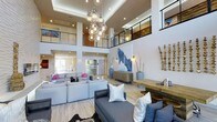 This sophisticated space makes an immediate impression with its dramatic double-height ceilings and open, airy layout. A striking cluster chandelier composed of glass globe pendants serves as a central focal point, drawing the eye upward and anchoring the expansive main living area. Light hardwood flooring runs throughout, contributing to a warm yet modern aesthetic.

The main gathering area is generously proportioned, featuring ample seating with plush sofas in soft grey and blue tones, accented by vibrant pink and white throw pillows that add a lively pop of color. A bold blue abstract artwork commands attention on one wall, complemented by sculptural décor pieces and tall vase arrangements that add organic texture to the space.

An upper mezzanine level, framed by sleek black metal railings, overlooks the main floor below, adding architectural depth and a sense of vertical grandeur. The space is bathed in natural and recessed lighting, enhancing the brightness and openness of the interior.

A console area with a woven-texture cabinet and decorative accessories provides a stylish transition point within the space. The overall design balances contemporary elegance with approachable comfort, making it a versatile and visually compelling environment suited for gathering, relaxing, or entertaining.