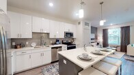 This single-floor unit presents a well-designed open-concept layout that seamlessly connects the kitchen, dining, and living areas. The kitchen is a standout feature, outfitted with crisp white cabinetry extending to the ceiling, a grey mosaic tile backsplash, and a full suite of stainless steel appliances including a range, microwave, and dishwasher. A central island with a white quartz countertop provides additional prep space and casual dining for multiple guests, illuminated by modern pendant lighting overhead. Wood-style flooring runs throughout the main living areas, adding warmth to the contemporary aesthetic.

The bedrooms are comfortably scaled, each benefiting from natural light through windows dressed with teal curtains and blinds. One bedroom features carpeted flooring and a ceiling fan, while another offers hardwood-style floors and a large floor mirror. The full bathroom is finished with white cabinetry, quartz countertops, wood-style tile flooring, and a full tub-shower combination. The overall ambiance of the unit is clean, modern, and cohesive, with a neutral color palette, recessed lighting, and consistent contemporary finishes carried throughout every room.