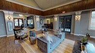 This thoughtfully designed single-floor communal space exudes a sophisticated yet comfortable ambiance, blending warm wood tones with contemporary styling throughout. The main lounge area commands attention with its dramatic vaulted ceilings and an expansive wall of floor-to-ceiling windows fitted with wood-slatted blinds, flooding the interior with generous natural light and offering views of the surrounding outdoor greenery. Rich wood-paneled upper walls complement the durable wood-look tile flooring, creating a cohesive and inviting aesthetic.

The open-concept layout features multiple seating arrangements, including plush sofas and accent chairs positioned atop layered area rugs, making the space ideal for both relaxation and social gatherings. Decorative columns with crown molding and wall sconces add architectural interest and a refined touch. A cozy bistro-style seating nook is tucked near the windows, providing an intimate spot for smaller gatherings.

Adjacent to the main lounge is a well-appointed kitchenette area, featuring a granite-topped bar counter with tufted leather barstools, gray shaker-style cabinetry, and full appliances set against a bold deep-teal accent wall. A glass-paned door leads to an additional room, further expanding the functional layout of this versatile and elegantly appointed space.