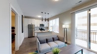 Located at 223 E Town St in the heart of Columbus, Ohio, this modern single-floor unit offers a thoughtfully designed open-concept layout that seamlessly integrates living and kitchen spaces. The kitchen is outfitted with stainless steel appliances, including a full-size refrigerator, microwave, and range, all complemented by crisp white cabinetry and a peninsula counter with bar seating. Sleek pendant lighting hangs above the peninsula, adding a contemporary touch to the space. The living area flows naturally from the kitchen, creating an inviting and functional environment. Hardwood-style flooring runs throughout, lending warmth and visual continuity to the space. A large window floods the interior with natural light while framing views of the surrounding Columbus cityscape. The neutral color palette of warm beige walls and white trim provides a clean, modern backdrop that suits a variety of interior styles. Vertical blinds offer privacy without compromising the light and openness of the unit. Overall, the space strikes a balance between modern design and everyday functionality, making it well-suited for urban living in downtown Columbus.