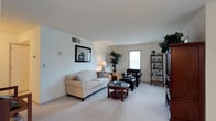 This single-floor space showcases a welcoming living room designed with a clean, neutral aesthetic. The room features light-colored walls and plush wall-to-wall carpeting in a warm beige tone, contributing to an overall sense of comfort and openness. Natural light filters in through a centrally positioned window, brightening the space without overwhelming it.

The living area is well-proportioned, offering ample room for a full seating arrangement. A cream-toned sofa anchors the space, complemented by a dark leather armchair and ottoman, creating a balanced contrast between light and dark furnishings. A round wooden coffee table with a glass top serves as a focal point at the center of the seating area.

Decorative elements add character throughout the room. A framed mirror mounted on one wall enhances the sense of depth, while a tall indoor plant near the window introduces a natural, organic touch. A dark wood shelving unit along one wall provides additional display and storage space. Overhead, a smooth, flat ceiling with a visible HVAC vent reflects a straightforward, functional design. The overall ambiance is tidy, understated, and move-in ready.