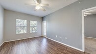 This efficient single-floor unit offers a well-organized layout comprising a combined kitchen and living area, one full bathroom, and a dedicated walk-in closet. Throughout the main living spaces, rich wood-look vinyl plank flooring adds warmth and durability, while soft gray walls create a clean, neutral backdrop.

The open living area is a standout feature, benefiting from two large windows that frame lush greenery outside and flood the room with natural light. A ceiling fan overhead enhances comfort, and the space feels open and airy thanks to the generous ceiling height. A glass-paneled door provides an additional point of natural light and likely leads to an outdoor area.

The kitchen is efficiently designed with white cabinetry offering ample storage, light-toned countertops, and a full suite of black appliances including an electric range, refrigerator, dishwasher, and built-in microwave. The kitchen flows seamlessly into the living space via an open doorway, maintaining a connected feel throughout.

The bathroom features wood-look flooring, white cabinetry, and a tiled shower-tub combination with a classic white tile surround accented by a golden tile border. The walk-in closet is carpeted and outfitted with wire shelving systems, providing organized storage. Overall, the unit presents a functional and tidy living environment with consistent finishes throughout.