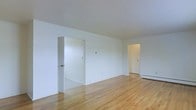 This single-floor space presents a straightforward and functional layout characterized by clean lines and a neutral aesthetic. The main living area is finished with warm-toned hardwood floors that run throughout, adding a classic and timeless quality to the interior. Crisp white walls and a white ceiling create a bright, airy atmosphere that feels open and uncluttered.

The room features multiple doorways that connect to adjacent spaces, providing clear circulation flow throughout the unit. A baseboard heating unit is visible along the far wall, indicating a practical and efficient heating setup well-suited for year-round comfort. The layout appears well-proportioned, offering enough space for a variety of furniture arrangements.

The overall ambiance is minimal and move-in ready, with a blank-canvas quality that allows for easy personalization. The combination of light-reflecting white surfaces and natural wood flooring gives the space a clean, welcoming feel. Whether used as a primary living area or repurposed for another function, this space offers a solid foundation with classic interior finishes and a simple, practical design throughout.