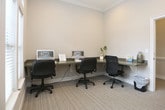 This single-floor space is thoughtfully designed as a functional business center, offering a quiet and professional environment well-suited for focused work. The layout features built-in wall-mounted desks with ample surface area, accommodating multiple workstations complete with computers and ergonomic seating. The neutral, warm-toned walls are complemented by clean white crown molding and baseboards, lending the space a polished and refined aesthetic. Carpet flooring in a subtle woven pattern adds warmth underfoot while contributing to a calm, noise-reduced atmosphere. Natural light filters in generously through large windows fitted with white horizontal blinds, allowing occupants to control light levels as needed. Views of lush greenery outside enhance the sense of openness. The entry is marked by a classic white paneled door with matte black hardware, providing a crisp contrast to the neutral interior palette. Recessed ceiling lighting ensures even illumination throughout. The space is efficiently organized, with each workstation area feeling distinct yet connected, making it ideal for shared use. Overall, the business center strikes a balance between practicality and comfort, creating an inviting setting for productivity.