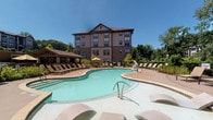 Located at 1731 Tupelo Hill Ln in Raleigh, NC, this outdoor amenity space is designed to offer a resort-style experience with a variety of well-appointed gathering areas. The centerpiece is a generously sized, freeform swimming pool surrounded by a broad sundeck lined with lounge chairs, ideal for relaxing under the North Carolina sun. Adjacent to the pool, a spacious covered pavilion features a striking vaulted timber-beam ceiling, ceiling fans, and stone-clad columns that provide a shaded retreat for outdoor dining and socializing. The pavilion accommodates multiple seating arrangements with wicker and metal dining sets, offering a comfortable al fresco environment. A built-in outdoor kitchen runs along a stone-faced wall and is equipped with multiple grills, making the area well-suited for communal gatherings. Beyond the covered area, an open patio extends the entertaining space and includes a fire pit with Adirondack-style seating, perfect for cooler evenings. Lush landscaping with flowering plants, manicured shrubs, and mature trees frames the entire outdoor area, enhancing privacy and creating a serene natural backdrop. Stone retaining walls and decorative planters add architectural cohesion throughout, giving the space a polished, upscale aesthetic.