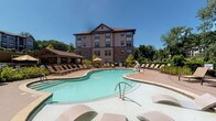 Located at 1731 Tupelo Hill Ln in Raleigh, NC, this outdoor amenity space is designed to offer a resort-style experience with a variety of well-appointed gathering areas. The centerpiece is a generously sized, freeform swimming pool surrounded by a broad sundeck lined with lounge chairs, ideal for relaxing under the North Carolina sun. Adjacent to the pool, a spacious covered pavilion features a striking vaulted timber-beam ceiling, ceiling fans, and stone-clad columns that provide a shaded retreat for outdoor dining and socializing. The pavilion accommodates multiple seating arrangements with wicker and metal dining sets, offering a comfortable al fresco environment. A built-in outdoor kitchen runs along a stone-faced wall and is equipped with multiple grills, making the area well-suited for communal gatherings. Beyond the covered area, an open patio extends the entertaining space and includes a fire pit with Adirondack-style seating, perfect for cooler evenings. Lush landscaping with flowering plants, manicured shrubs, and mature trees frames the entire outdoor area, enhancing privacy and creating a serene natural backdrop. Stone retaining walls and decorative planters add architectural cohesion throughout, giving the space a polished, upscale aesthetic.