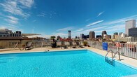 This rooftop pool deck in New Orleans, LA, offers a dynamic outdoor and indoor experience set against a sweeping panoramic view of the city's iconic skyline. The outdoor deck centers around a generously sized swimming pool with vivid turquoise water, surrounded by a light-toned deck surface. A collection of lounge chairs and a shaded umbrella provide comfortable spots for sunbathing, while potted palms and tropical greenery add a resort-like ambiance. A railed walkway encircles the deck, maximizing unobstructed sightlines to the downtown skyline under open skies.

Sliding glass doors connect the outdoor deck to an interior lounge area, distinguished by its bold color palette—yellow upper walls contrast with blue-gray lower walls adorned with large-scale aquatic-themed artwork. The polished concrete flooring in deep blue-gray tones anchors the space, which is furnished with a sectional sofa, accent chairs in orange cushions, and a central coffee table. A service bar area with reflective tile backsplash and wall-mounted television adds functionality. An adjacent dining zone features a tall wooden table with high-back chairs, positioned near sliding glass doors that frame additional skyline views and provide seamless indoor-outdoor flow.