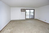 This single-floor space offers a functional and straightforward layout designed for comfortable everyday living. The main living area is finished with wall-to-wall carpeting that adds warmth underfoot, while smooth white walls provide a clean, neutral backdrop throughout. A large, multi-pane window allows generous natural light to pour into the room, creating a bright and open atmosphere. Climate comfort is well-addressed with a wall-mounted air conditioning unit positioned near the window for efficient cooling, complemented by baseboard heating that runs along the perimeter of the room for consistent warmth during cooler months. The textured ceiling adds a subtle architectural detail consistent with the space's practical character. The overall ambiance is understated and move-in ready, offering a blank canvas that can be easily personalized. The open feel of the main room provides flexibility in furniture arrangement, making it adaptable to a variety of uses. This space is well-suited for those seeking a no-frills, functional environment with essential amenities already in place.