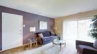 This single-floor residence offers a welcoming and thoughtfully designed living space that balances comfort with style. The living room serves as the heart of the home, immediately drawing attention with its striking deep purple accent wall that adds a bold splash of color against the surrounding neutral, warm-toned walls. Soft, plush carpeting underfoot enhances the cozy atmosphere, making the space feel inviting and comfortable.

Natural light plays a central role in the room's ambiance, pouring in generously through a large sliding glass door dressed with bright vertical blinds. This feature not only brightens the interior but also provides a seamless visual connection to the outdoors. The room accommodates a classic sofa paired with coordinating glass-top side and coffee tables, creating a functional yet elegant seating arrangement. Ambient lighting from stylish table lamps on either side of the sofa adds warmth during evening hours.

A framed piece of artwork anchors the accent wall, adding a finishing touch to the living area's decor. The overall design of this space reflects a clean, comfortable aesthetic with attention to detail, making it a practical and pleasant place to relax and entertain.