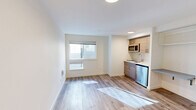 Located at 4629 21st Ave NE in Seattle's University District, this studio unit offers a thoughtfully designed, efficient layout that maximizes every square foot of its compact footprint. The open living area is finished with light-toned wood-look flooring throughout, crisp white walls, and recessed flush-mount lighting, creating a clean, contemporary aesthetic that feels bright and welcoming.

The kitchenette is neatly tucked along one wall and features warm wood-grain cabinetry paired with a white countertop, an undermount sink, a stainless steel compact refrigerator, and an over-the-range microwave — providing all essential cooking and storage amenities in a space-conscious design. Adjacent to the kitchen, a built-in shelving nook offers convenient additional storage or workspace potential.

The bathroom is accessible through a dedicated door and includes a toilet, sink with mirror, and a glass sliding-door shower enclosure with brushed nickel hardware. A built-in open closet with shelving and hanging rods is positioned just outside the bathroom, offering organized storage within the main living space.

A window along the main living wall draws in natural light, while the entry door area includes a thermostat panel and ventilation unit. The overall design is modern, clean, and efficiently arranged, making this unit a practical option in the heart of Seattle.