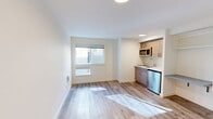 Located at 4629 21st Ave NE in Seattle's University District, this studio unit offers a thoughtfully designed, efficient layout that maximizes every square foot of its compact footprint. The open living area is finished with light-toned wood-look flooring throughout, crisp white walls, and recessed flush-mount lighting, creating a clean, contemporary aesthetic that feels bright and welcoming.

The kitchenette is neatly tucked along one wall and features warm wood-grain cabinetry paired with a white countertop, an undermount sink, a stainless steel compact refrigerator, and an over-the-range microwave — providing all essential cooking and storage amenities in a space-conscious design. Adjacent to the kitchen, a built-in shelving nook offers convenient additional storage or workspace potential.

The bathroom is accessible through a dedicated door and includes a toilet, sink with mirror, and a glass sliding-door shower enclosure with brushed nickel hardware. A built-in open closet with shelving and hanging rods is positioned just outside the bathroom, offering organized storage within the main living space.

A window along the main living wall draws in natural light, while the entry door area includes a thermostat panel and ventilation unit. The overall design is modern, clean, and efficiently arranged, making this unit a practical option in the heart of Seattle.