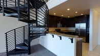 This two-floor space makes an immediate impression with its dramatic design choices and cohesive aesthetic. At the heart of the main level, a bold black spiral staircase serves as both a functional element and a striking architectural focal point. Its open metalwork railings and sweeping form draw the eye upward, connecting the two floors with visual flair.

The kitchen is thoughtfully appointed and features rich, dark cabinetry that extends to the ceiling, providing ample storage while reinforcing the space's sophisticated color palette. Granite countertops with warm, speckled tones offer a refined contrast to the dark cabinetry and are complemented by a peninsula-style breakfast bar with corbel supports — an inviting spot for casual dining or entertaining. Stainless steel appliances, including a microwave and refrigerator, add a modern, polished finish to the culinary space.

Recessed lighting throughout the kitchen and living area provides warm, even illumination, enhancing the overall ambiance. The flooring features plush carpeting that adds comfort underfoot, while the crisp white walls create a clean backdrop that allows the dark design elements to stand out. The layout flows naturally between areas, making the space feel open and well-organized across both levels.