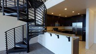 This two-floor space makes an immediate impression with its dramatic design choices and cohesive aesthetic. At the heart of the main level, a bold black spiral staircase serves as both a functional element and a striking architectural focal point. Its open metalwork railings and sweeping form draw the eye upward, connecting the two floors with visual flair.

The kitchen is thoughtfully appointed and features rich, dark cabinetry that extends to the ceiling, providing ample storage while reinforcing the space's sophisticated color palette. Granite countertops with warm, speckled tones offer a refined contrast to the dark cabinetry and are complemented by a peninsula-style breakfast bar with corbel supports — an inviting spot for casual dining or entertaining. Stainless steel appliances, including a microwave and refrigerator, add a modern, polished finish to the culinary space.

Recessed lighting throughout the kitchen and living area provides warm, even illumination, enhancing the overall ambiance. The flooring features plush carpeting that adds comfort underfoot, while the crisp white walls create a clean backdrop that allows the dark design elements to stand out. The layout flows naturally between areas, making the space feel open and well-organized across both levels.
