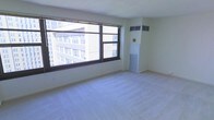 This single-floor space offers a clean, minimalist environment with a bright and open feel. The main living area is defined by expansive windows with dark frames that flood the room with natural light and frame striking views of the surrounding urban cityscape, with multi-story buildings visible just beyond the glass. The light-colored carpeting throughout contributes to the airy, neutral aesthetic of the interior. The walls are finished in a crisp, off-white tone, giving the space a fresh and versatile canvas. A wall-mounted HVAC unit and a built-in floor-level air return provide year-round climate control, seamlessly integrated into the room's design. The layout is open and unobstructed, allowing for flexible furniture arrangement and use of the available floor space. The overall ambiance is calm and functional, with an urban energy brought in through the generous window presence. The space is well-suited for a range of purposes, benefiting from its elevated city views, abundant natural light, and straightforward, practical interior design.