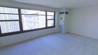 This single-floor space offers a clean, minimalist environment with a bright and open feel. The main living area is defined by expansive windows with dark frames that flood the room with natural light and frame striking views of the surrounding urban cityscape, with multi-story buildings visible just beyond the glass. The light-colored carpeting throughout contributes to the airy, neutral aesthetic of the interior. The walls are finished in a crisp, off-white tone, giving the space a fresh and versatile canvas. A wall-mounted HVAC unit and a built-in floor-level air return provide year-round climate control, seamlessly integrated into the room's design. The layout is open and unobstructed, allowing for flexible furniture arrangement and use of the available floor space. The overall ambiance is calm and functional, with an urban energy brought in through the generous window presence. The space is well-suited for a range of purposes, benefiting from its elevated city views, abundant natural light, and straightforward, practical interior design.