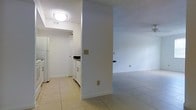 This single-floor space presents a clean, straightforward layout connecting a functional kitchen to an open living area. The kitchen is finished with white cabinetry, white appliances including a refrigerator and range, and dark countertops, creating a crisp, utilitarian aesthetic. A louvered pantry or closet door adds practical storage within the kitchen zone. Light ceramic tile flooring runs consistently throughout, lending a cohesive and easy-to-maintain surface underfoot. The walls are painted a bright white, contributing to an airy, well-lit atmosphere. A flush-mount ceiling light provides overhead illumination in the kitchen area, while a textured ceiling adds a subtle visual detail. A central structural column or partial wall separates the kitchen from the adjoining living area, which features the same tile flooring and neutral white walls. The living space feels open and versatile, offering a blank canvas for various furniture arrangements. Electrical outlets and switches are conveniently positioned throughout. Overall, the space maintains a clean, minimal character that is functional and easy to personalize.