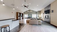 Located in West Valley City, Utah, this well-appointed single-floor community space offers an open and airy layout designed for comfort and functionality. Soaring vaulted ceilings with recessed lighting and ceiling fans lend a sense of grandeur throughout, while large windows flood the interior with natural light and offer views of the surrounding greenery.

The kitchen is a standout feature, boasting crisp white shaker cabinetry, dark granite countertops, stainless steel appliances including a full-size refrigerator, and a central island with a sink. A bar-height counter with three industrial-style stools provides casual seating, while pendant lights add a warm, modern accent overhead. Dark tile flooring grounds the kitchen area with a rich, durable finish.

The adjoining living room centers around a dramatic floor-to-ceiling stone fireplace with a mounted television, complemented by contemporary seating and light wood-plank flooring. The dining area nearby accommodates a large rectangular table with seating for multiple guests, framed by patterned drapes and vibrant wall art.

A flexible open area near the entrance, accented by textured wall coverings, hanging planters, and warm-toned greenery, provides an inviting transition into the space. Dark wood double doors with glass panels complete the welcoming entryway aesthetic.