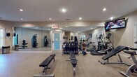 This fitness center is a comprehensively outfitted exercise facility designed to support a wide range of workout routines. The space is finished with durable speckled flooring throughout and illuminated by recessed ceiling lighting, creating a clean, functional atmosphere.

The cardio zone features multiple treadmills and an elliptical trainer lined along a windowed wall fitted with classic plantation shutters, allowing natural light to filter in. A wall-mounted flat-screen TV is positioned for easy viewing during workouts, adding an entertainment element to cardio sessions.

The free weights area includes a full dumbbell rack stocked with a comprehensive range of weights, accompanied by adjustable workout benches and an assortment of medicine balls, catering to both strength and functional training needs.

A dedicated strength training section houses a multi-use cable and resistance machine, with additional benches and weight equipment nearby. Floor-to-ceiling mirrors line multiple walls throughout the facility, visually expanding the space while allowing users to monitor their form.

The facility also includes a double-door storage closet, clearly posted fitness center rules and notices, and a dedicated exit door. The neutral, warm-toned walls and thoughtful layout of equipment throughout contribute to an organized and welcoming environment suitable for all fitness levels.