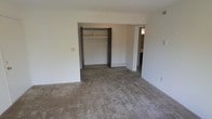 This single-floor space presents a straightforward and functional layout. The primary room shown is a generously sized bedroom, finished with wall-to-wall carpet in a neutral taupe tone that extends throughout the space. The room features plain white walls and a standard ceiling height, lending a clean, understated aesthetic.

A built-in closet is prominently positioned along the back wall, featuring a double-door opening with a shelf and hanging rod, providing practical storage. The closet opening is framed with dark brown trim that contrasts against the light walls. Adjacent to the closet, an open doorway leads to a connected room — likely a bathroom or hallway — visible in the background, suggesting a convenient en-suite or pass-through configuration.

A floor vent is visible near the base of the wall, indicating a forced-air HVAC system. Electrical outlets and a switch plate are positioned along the walls, and an additional vent can be noted near the ceiling. The overall ambiance is neutral and move-in ready, offering a blank canvas suitable for a variety of furnishing arrangements. The space is vacant, allowing for a clear view of the room's proportions and layout.