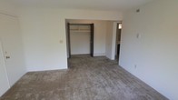 This single-floor space presents a straightforward and functional layout. The primary room shown is a generously sized bedroom, finished with wall-to-wall carpet in a neutral taupe tone that extends throughout the space. The room features plain white walls and a standard ceiling height, lending a clean, understated aesthetic.

A built-in closet is prominently positioned along the back wall, featuring a double-door opening with a shelf and hanging rod, providing practical storage. The closet opening is framed with dark brown trim that contrasts against the light walls. Adjacent to the closet, an open doorway leads to a connected room — likely a bathroom or hallway — visible in the background, suggesting a convenient en-suite or pass-through configuration.

A floor vent is visible near the base of the wall, indicating a forced-air HVAC system. Electrical outlets and a switch plate are positioned along the walls, and an additional vent can be noted near the ceiling. The overall ambiance is neutral and move-in ready, offering a blank canvas suitable for a variety of furnishing arrangements. The space is vacant, allowing for a clear view of the room's proportions and layout.