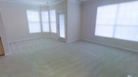 This single-floor space presents a spacious, open living area finished in a clean, neutral color palette. The room is carpeted in a light beige, wall-to-wall plush carpet that adds warmth underfoot and complements the soft, warm-toned walls. Crown molding runs along the ceiling perimeter, lending a refined architectural detail that elevates the overall aesthetic.

Natural light is a defining feature of this space, with multiple windows fitted with horizontal blinds positioned along the walls, including a charming angled bay-style window arrangement in one corner that draws in generous amounts of daylight from multiple directions. An additional window on the adjacent wall further enhances the bright, airy feel of the room.

A glass-paneled door provides both access and additional light, contributing to the open, welcoming atmosphere. The wide baseboards throughout the room add another layer of classic detailing consistent with the crown molding above.

Overall, the space offers a clean, move-in-ready environment with classic architectural touches, abundant natural light, and a neutral design foundation that accommodates a wide range of interior styles.