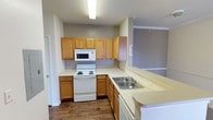 This single-floor residence offers a straightforward and functional layout designed for comfortable everyday living. The kitchen is well-equipped with warm oak cabinetry spanning both upper and lower sections, light-toned countertops, a built-in over-the-range microwave, a white electric coil range, and a double stainless steel sink. The L-shaped kitchen configuration provides ample counter and storage space, opening toward the main living area. The living area features neutral-toned carpet, crown molding, a ceiling fan, and large sliding glass doors dressed with vertical blinds that allow natural light to fill the space. Adjacent to the living area, a hallway leads to the bathroom, which includes a vanity with a sink, mirror, overhead wall cabinet for storage, and a toilet. The bedrooms are carpeted and outfitted with ceiling fans; one bedroom benefits from natural light through a pair of windows with blinds, while another features a more enclosed, private feel. Throughout the home, walls are painted in soft neutral tones, and crown molding accents several areas, adding a refined touch. The combination of hardwood-style flooring in the kitchen and transitional areas with carpet in the living and sleeping spaces creates a cohesive and practical interior.