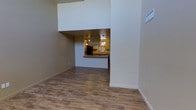 This single-floor residence showcases a thoughtfully designed open-concept layout that maximizes both space and functionality. The living area features warm, wood-look hardwood-style flooring that flows seamlessly throughout, complemented by neutral-toned walls that create a clean and inviting atmosphere. Notably high ceilings add a dramatic sense of volume and airiness to the main living space, enhancing the overall sense of openness.

The kitchen is a standout feature, visible through a pass-through opening that connects it to the main living area. It is outfitted with warm wood cabinetry, a black refrigerator, and a generous countertop breakfast bar that provides both additional workspace and a casual dining option. Pendant lighting hangs above the kitchen area, adding a warm, focused glow that accentuates the cabinetry and countertops. Window treatments in the kitchen allow for natural light control while maintaining a cozy ambiance.

Architectural details such as the bold upper wall cutout and clean-lined structural elements add visual interest and a modern sensibility to the space. The overall design balances practicality with style, making it a comfortable and well-organized living environment.