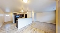 This single-floor space showcases a thoughtfully designed open-concept layout that seamlessly connects the kitchen and living areas. The kitchen is appointed with dark espresso-toned cabinetry, black appliances including a refrigerator, range, and overhead microwave, and a peninsula-style breakfast bar that serves as a natural divider between the kitchen and the main living space. Pendant lighting above the bar adds a warm, inviting ambiance, complemented by flush-mount ceiling fixtures throughout. The kitchen features smooth light-toned countertops that contrast elegantly with the dark cabinetry. The flooring transitions from wood-look planks in the kitchen and entryway to plush carpet in the living area, creating distinct zones within the open floor plan. Neutral gray walls and white trim provide a clean, versatile backdrop throughout the space. A hallway visible from the living area leads to additional rooms, with a paneled door offering privacy beyond. The overall atmosphere is modern yet cozy, with a practical layout well-suited for both everyday living and entertaining.